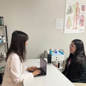 A healthcare professional in a white coat types on a laptop, while a patient with long hair sits on the other side of the desk. Medical posters and supplies are visible in the room.