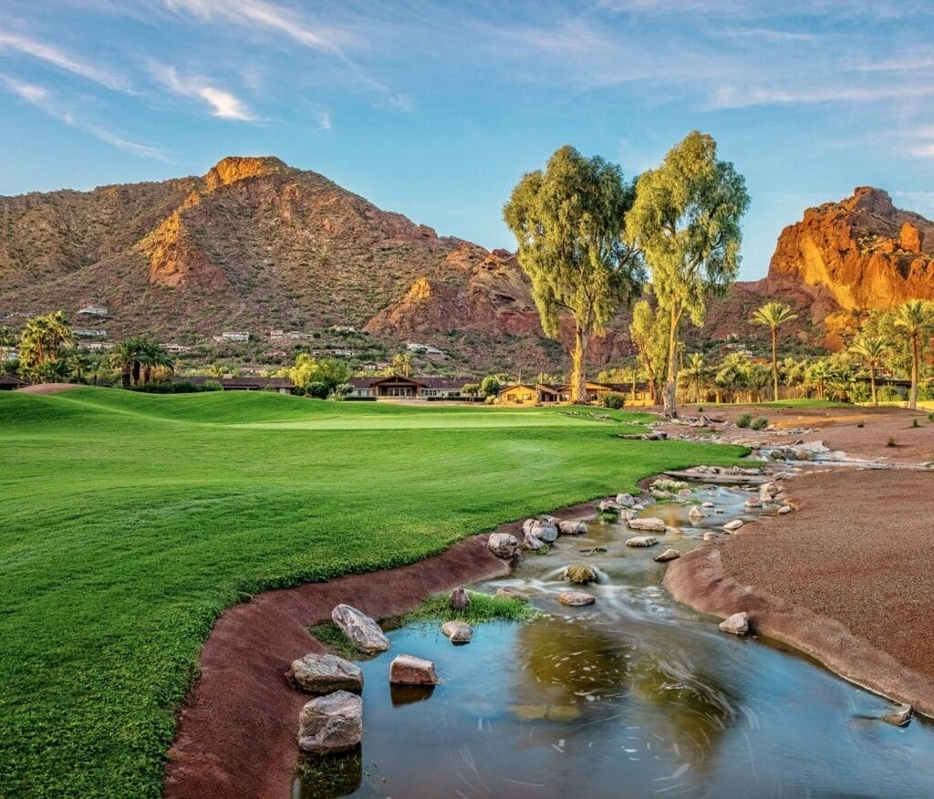 A scenic landscape with a green lawn, a small stream with stones, and desert mountains in the background under a clear blue sky.